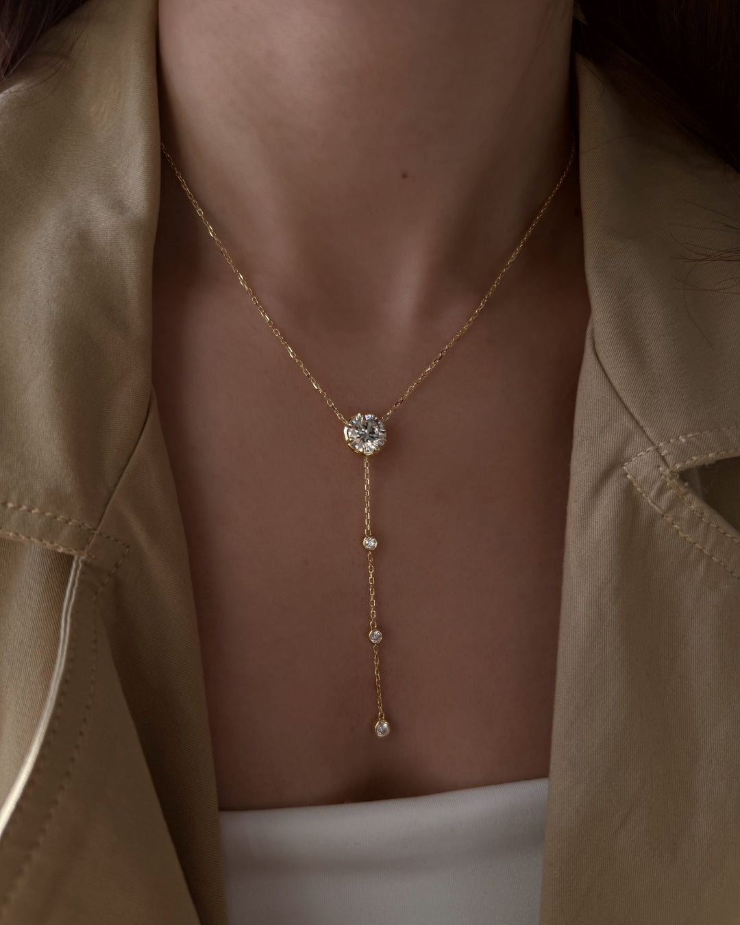 A close-up of a model wearing a delicate gold lariat necklace with a clear round-cut moissanite center stone and three small dangling gems, styled with a beige satin jacket.