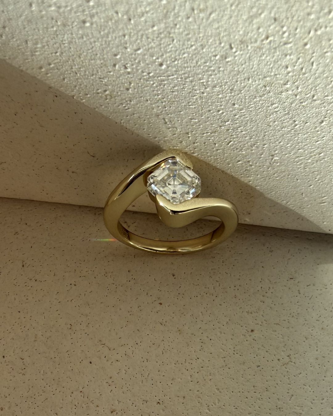 Gold asscher cut ring photographed in natural light on a textured surface highlighting its geometric stone