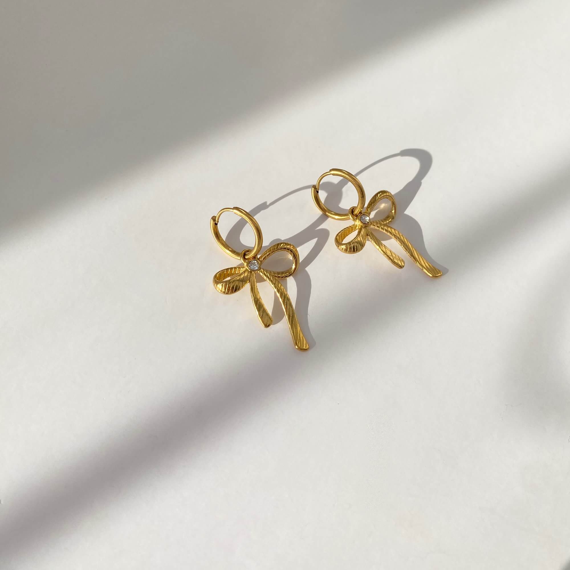 Angled flat lay of KrissaE dainty bow dangle earrings by Kampy Designs, showing shimmer and soft feminine detailing from the side.