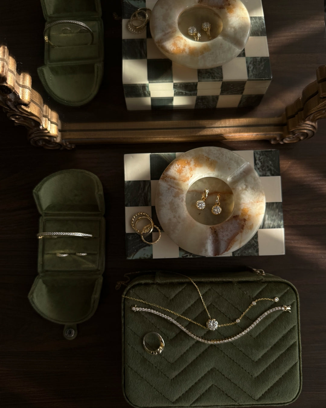 An overhead styled flat-lay of moissanite jewelry by Kampy Designs on a dark wood surface, including an open quilted olive green pouch with a lariat chain necklace draped across it, matching jewelry pieces, rings, and a checkered box in the background.