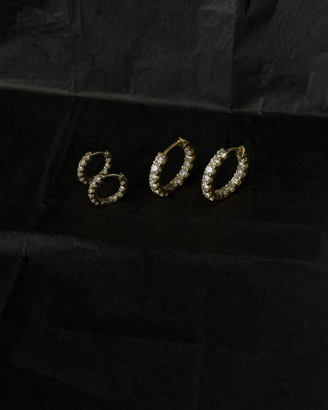 Gold moissanite hoop earrings displayed with smaller matching huggie hoops on a black textured background.
