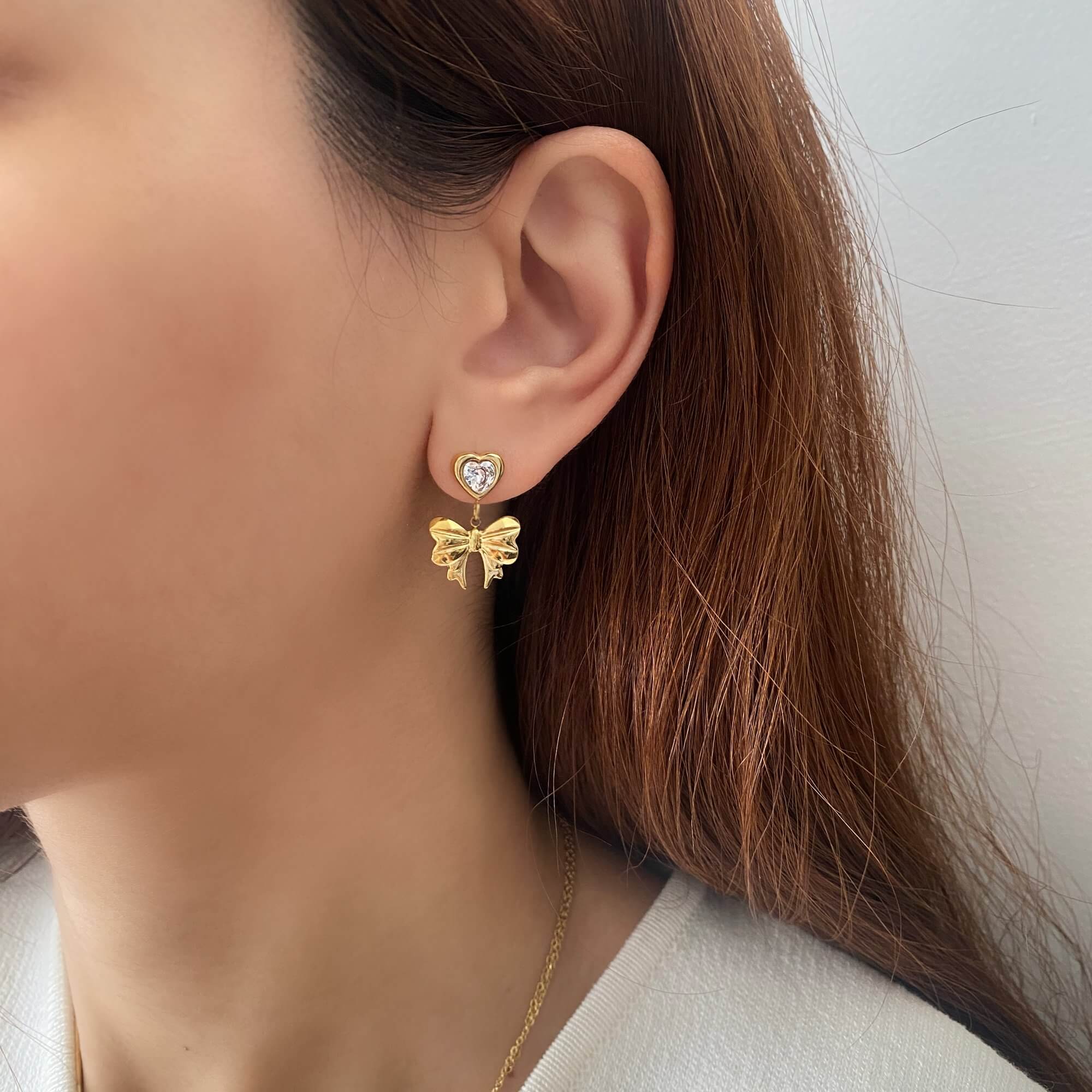 Close-up of KutieE heart bow dangle earrings by Kampy Designs, featuring a delicate bow above a dangling heart, styled on model's ear.