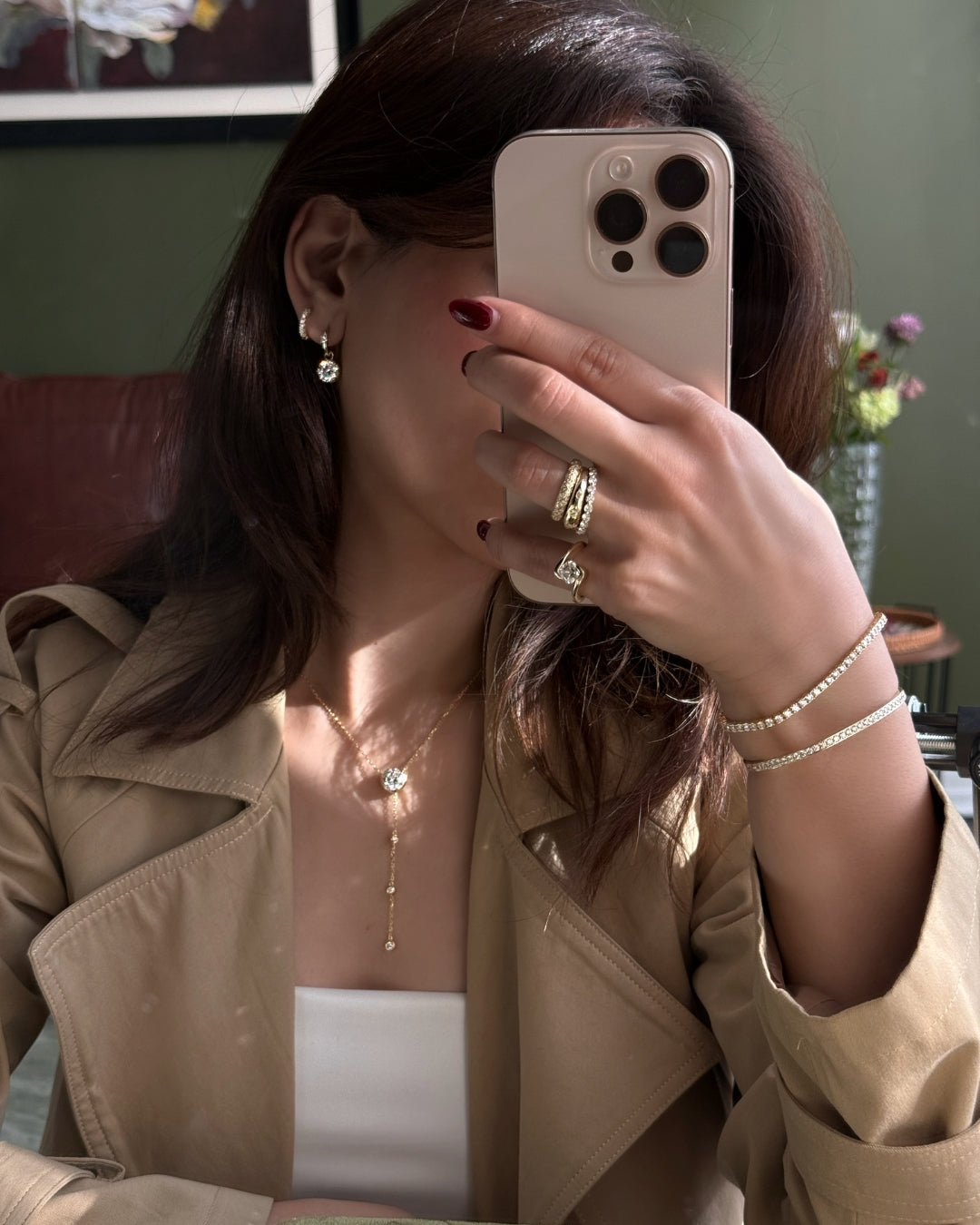 Model takes a mirror selfie showcasing gold jewelry by Kampy Designs, including a delicate gold lumina lariat necklace, a gold ring stack, and a tennis bracelet, while wearing a beige coat.