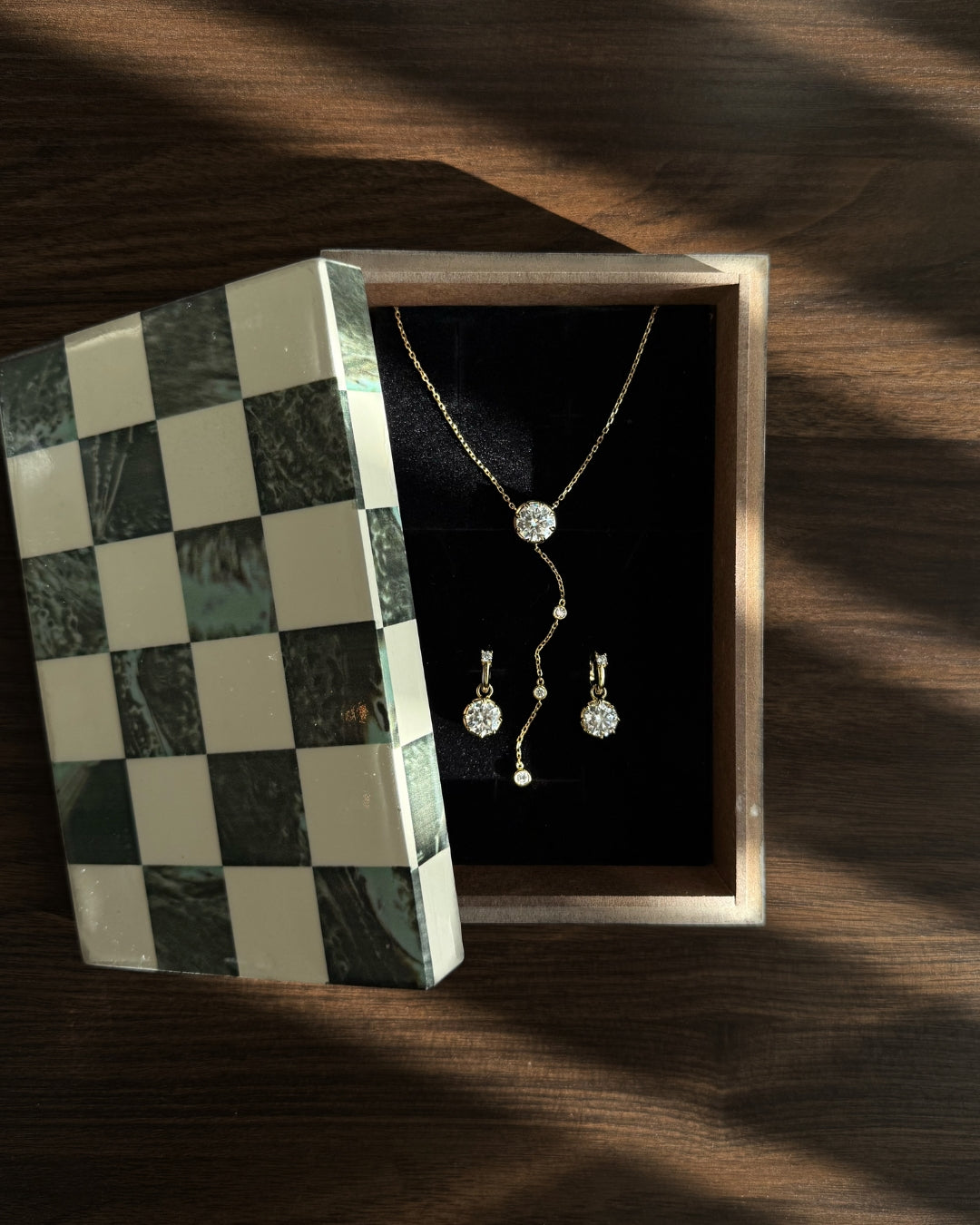 A lariat necklace and drop earrings jewelry gift set by Kampy Designs displayed inside an open wooden box with a green and cream checkered marble-patterned lid, set on a dark wooden surface with dramatic diagonal shadows.