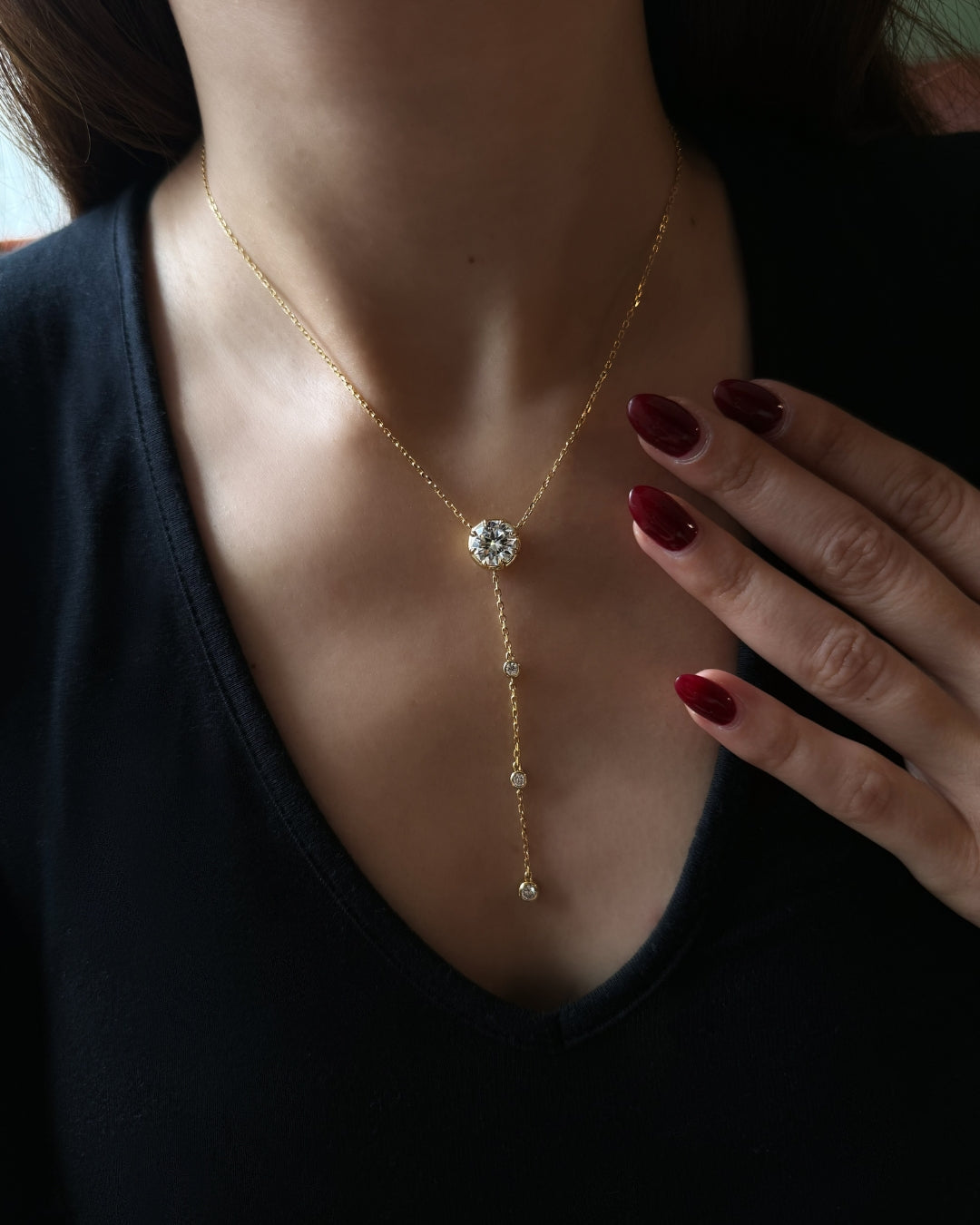 A close-up of a model wearing a delicate gold Y-necklace with a round clear moissanite gemstone centerpiece and small dangling gems, paired with a black top.