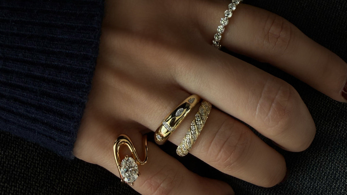 A close-up of a hand wearing multiple gold rings, including a moissanite-accented ring and a pavé band ring, set against a dark textured background.