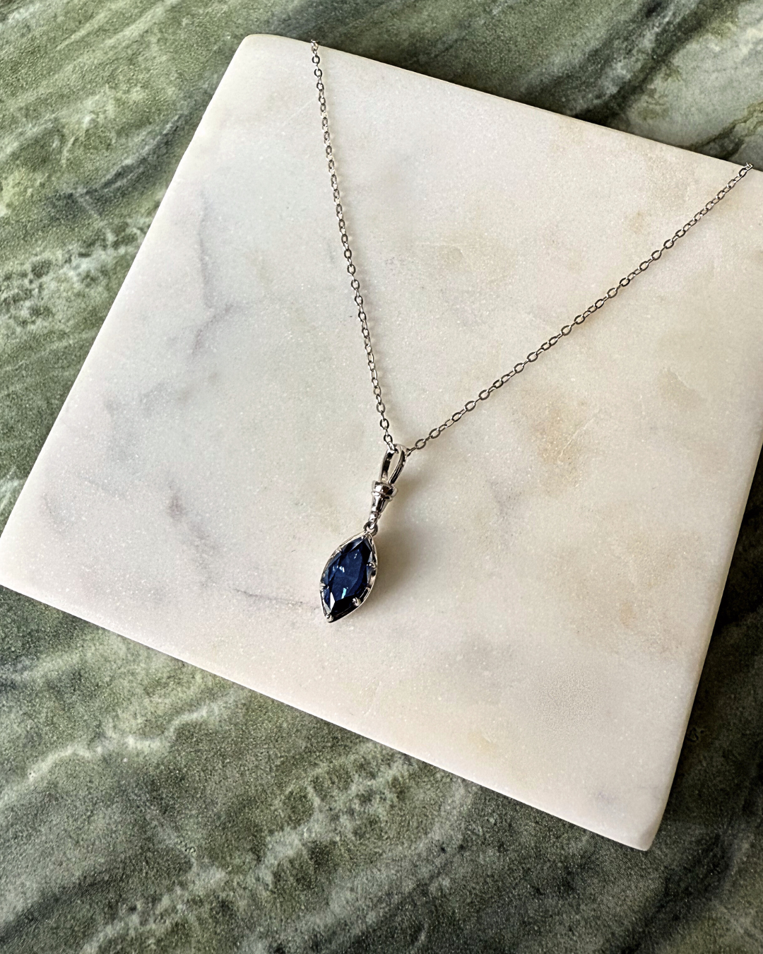 Marquise midnight blue moissanite pendant necklace displayed on marble surface with sterling silver chain.