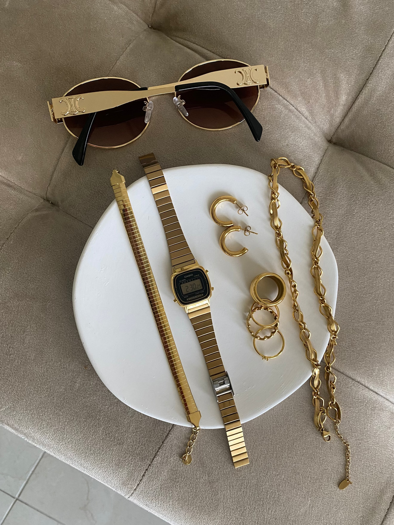 Gold sunglasses, watch, and jewelry on a white plate with a beige cushion background