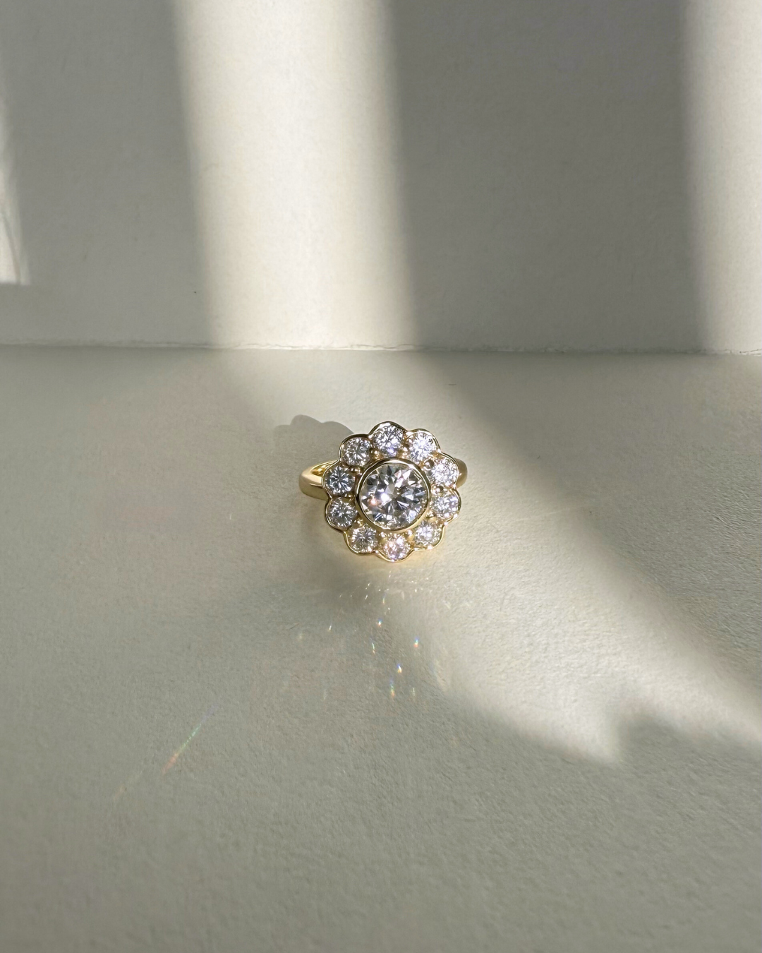 The Marigold Ring by Kampy Designs placed on a neutral background with sunlight beams, showing the round moissanite floral halo design in gold.