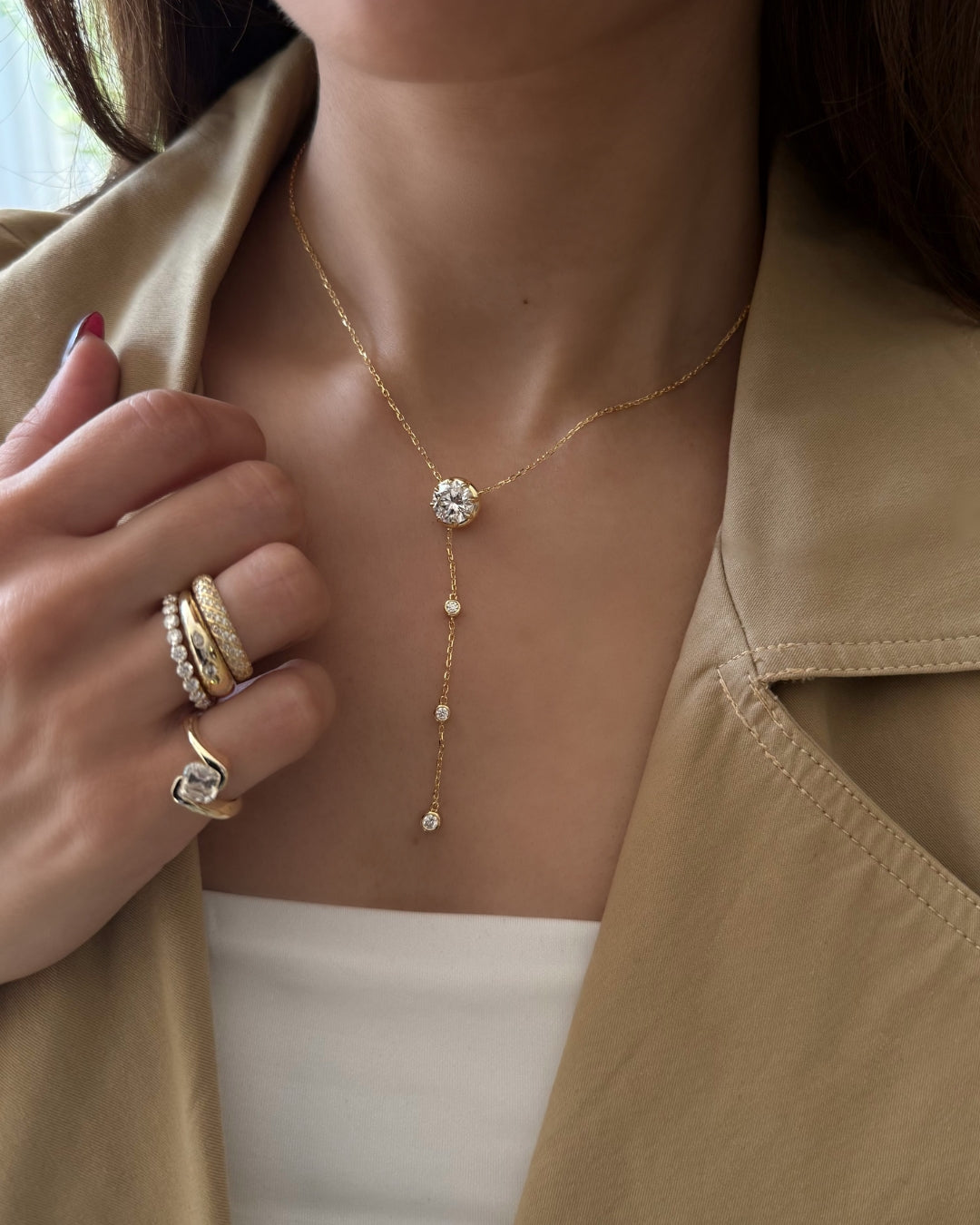 A close-up of a model wearing a delicate gold Y-necklace with a round 3 CT clear moissanite stone centerpiece and small bezel moissanites dangled, styled with gold rings and a beige blazer.