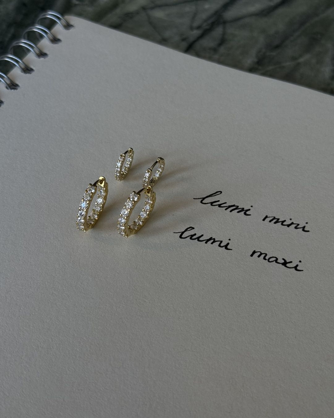 Gold moissanite huggie hoop earrings and mini hoops placed on a sketchbook with handwritten size labels.