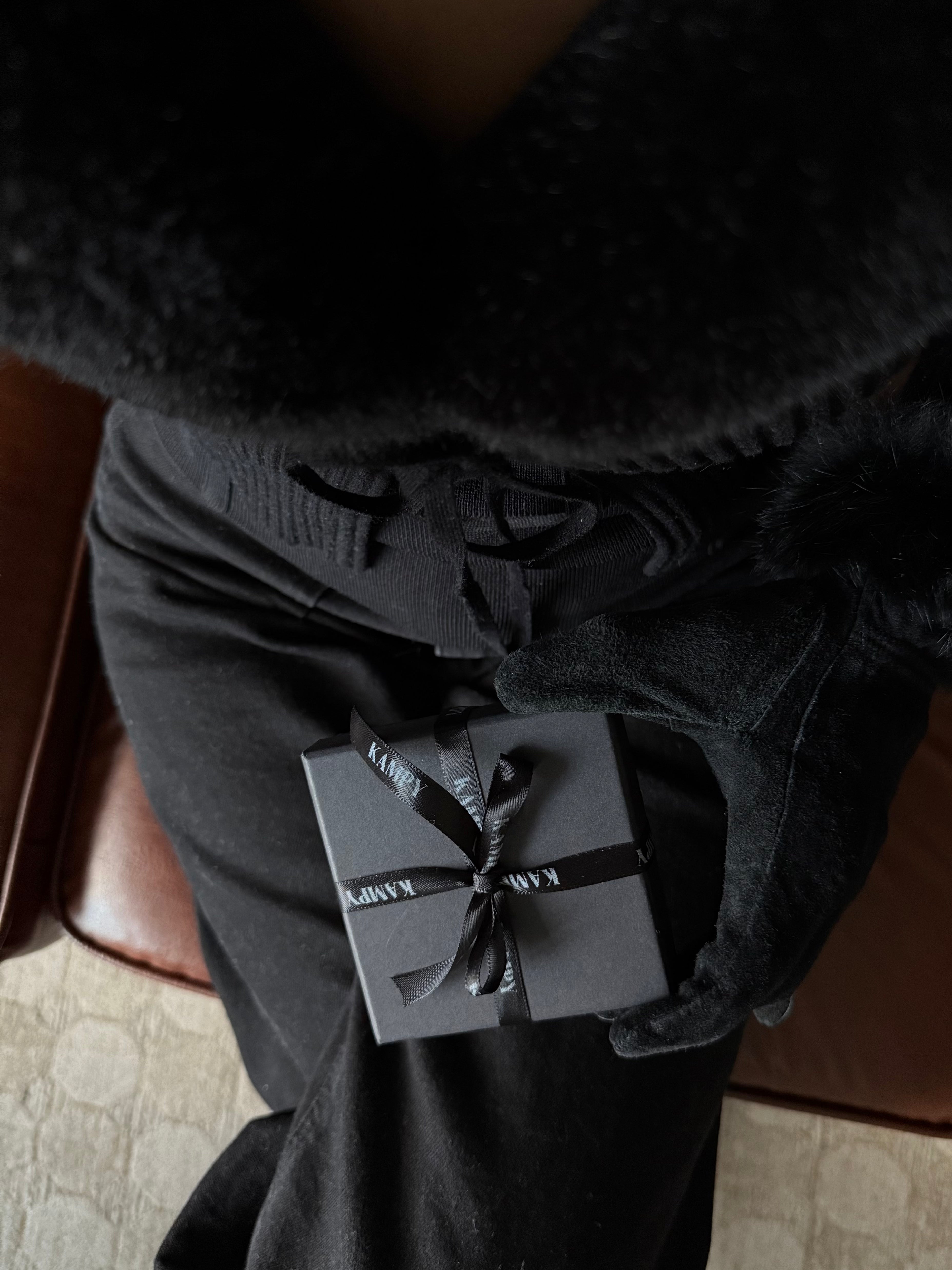 Person holding a small black gift box with a ribbon, wearing a dark coat.