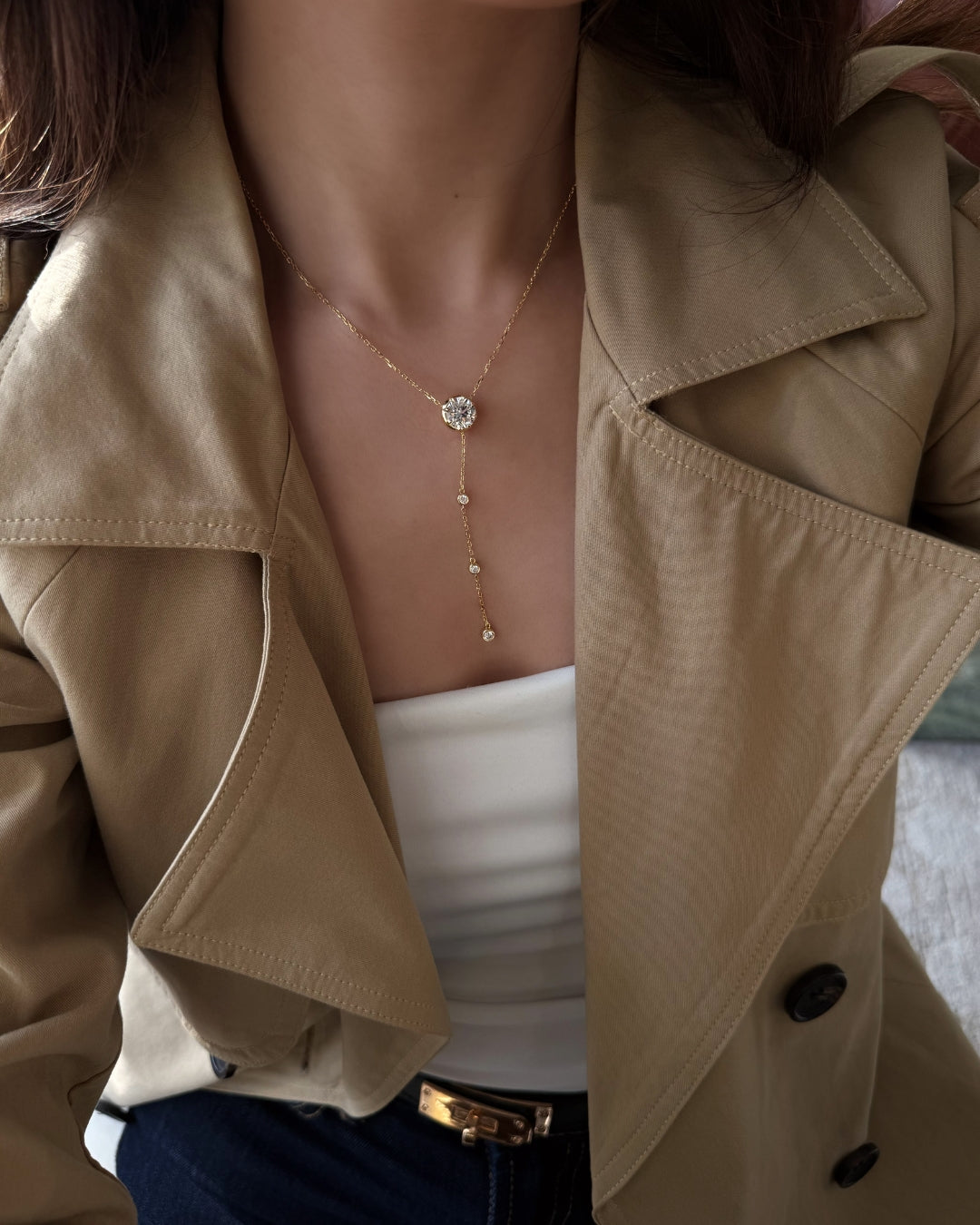 A close-up of a person wearing a gold lariat-style necklace with a round clear moissanite stone pendant and small dangling gems, styled with a beige trench coat and white top.