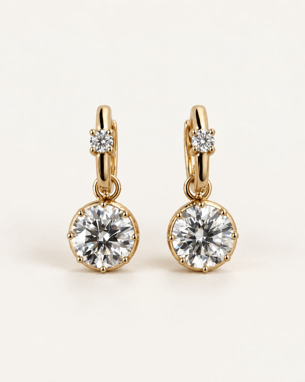 A pair of gold hoop earrings with a small round clear moissanite gemstone in the upper loop and a 2 carat round clear moissanite gemstone in a bezel setting hanging below, shown on a light cream background.
