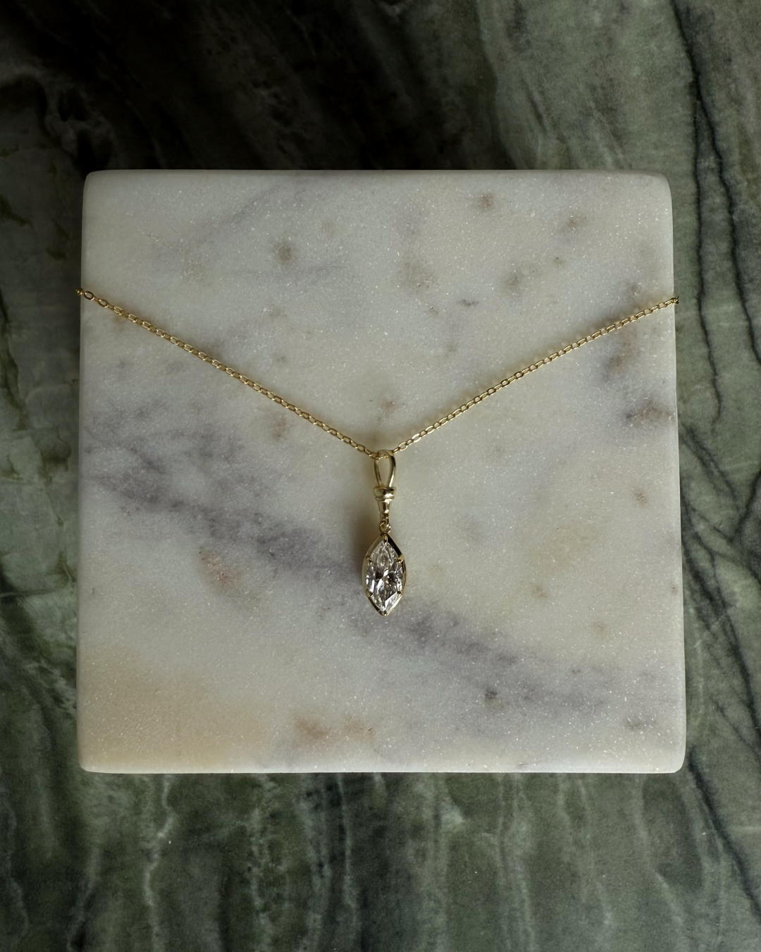 Flat lay of marquise white moissanite necklace with 2ct center stone on gold chain displayed on marble surface by Kampy