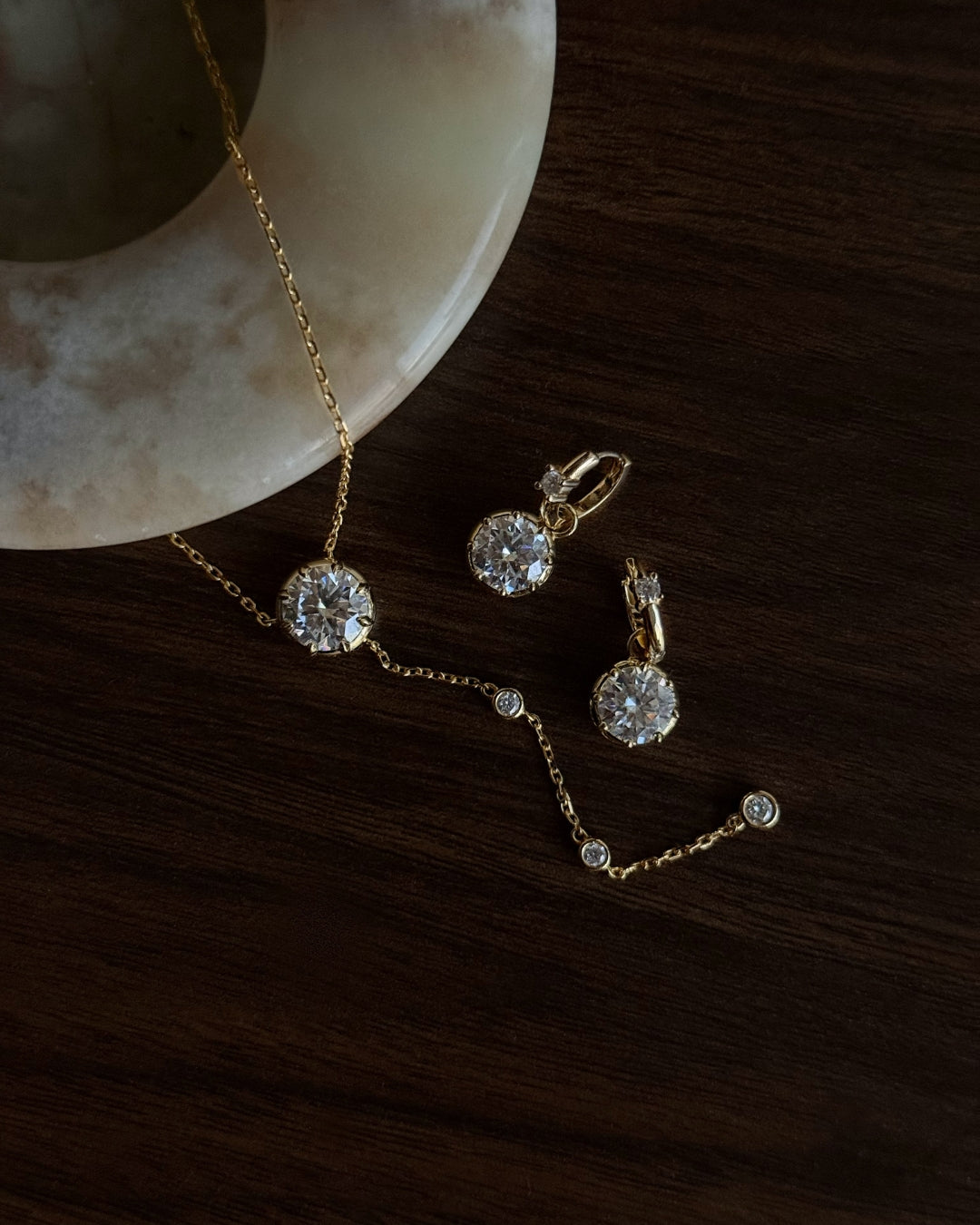 Gold jewelry set with a lariat necklace and drop earrings with round clear moissanite gemstones, arranged on a dark wood surface next to a cream-colored stone dish.